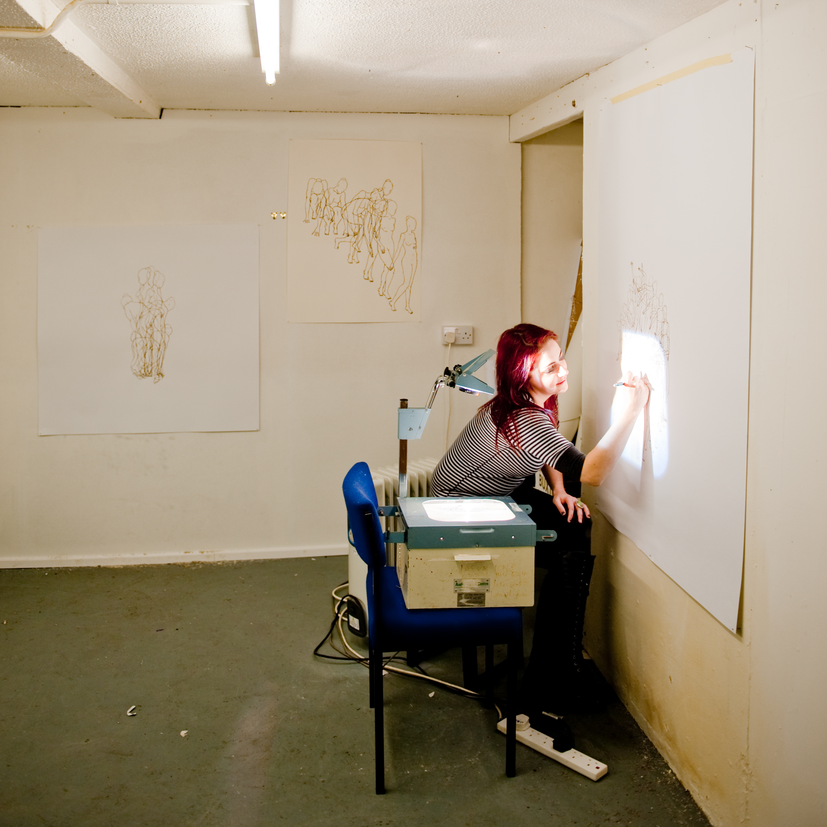 Photo of a woman sitting next to a projector, drawing onto paper attached to the wall